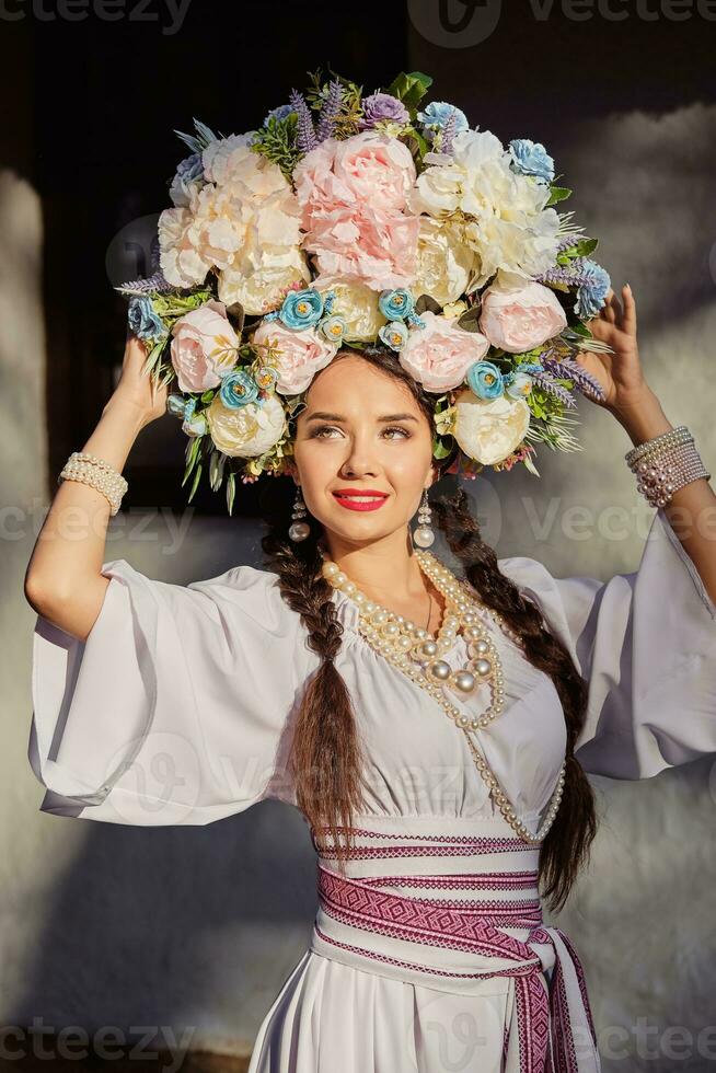 morena niña en un blanco ucranio auténtico nacional disfraz y un guirnalda de flores es posando en contra un blanco cabaña. de cerca. foto