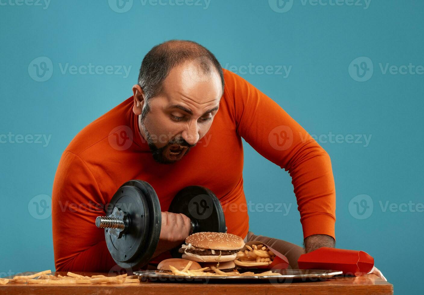 Close-up portrait of a middle-aged man with beard, dressed in a red turtleneck, posing with burgers and french fries. Blue background. Fast food. photo