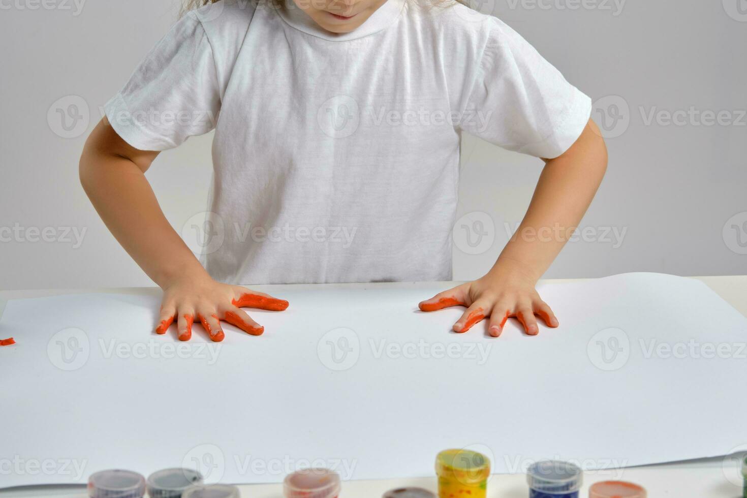 Little artist in white t-shirt standing at table with whatman and colorful paints, painting on it with her hands. Isolated on white. Close-up. photo