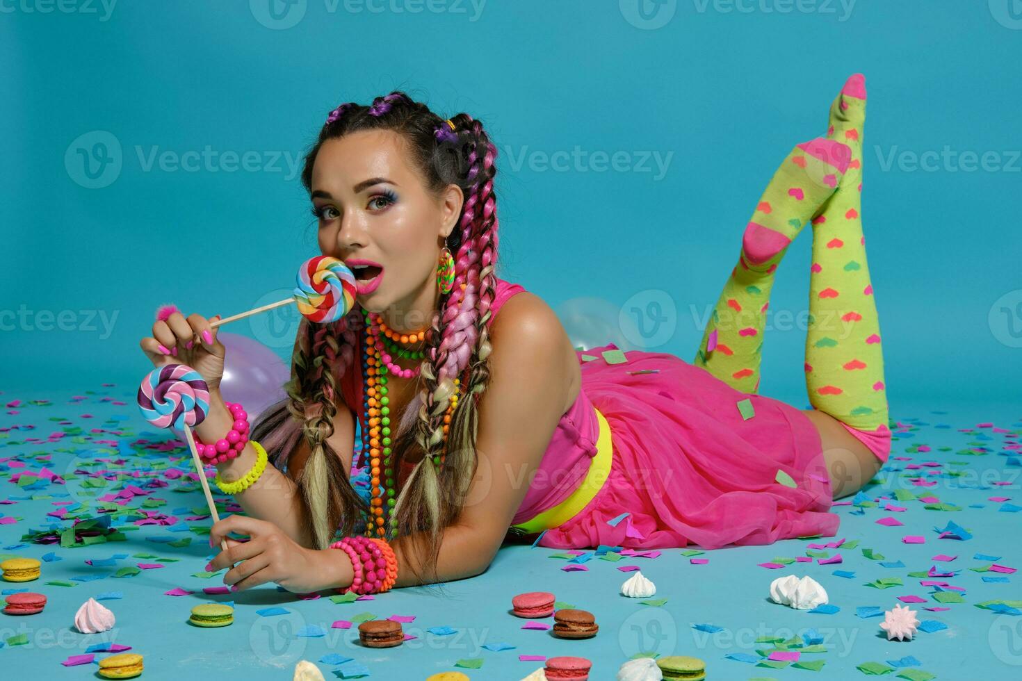 Lovely girl with a multi-colored braids hairstyle and bright make-up, posing in studio with lollipop, air balloons and confetti against a blue background. photo