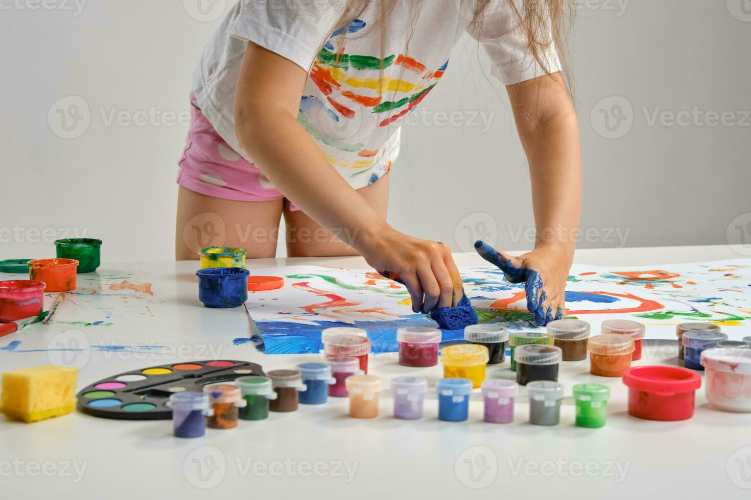 Little artist in white t-shirt standing at table with whatman and colorful paints, painting on it with her hands. Isolated on white. Close-up. photo