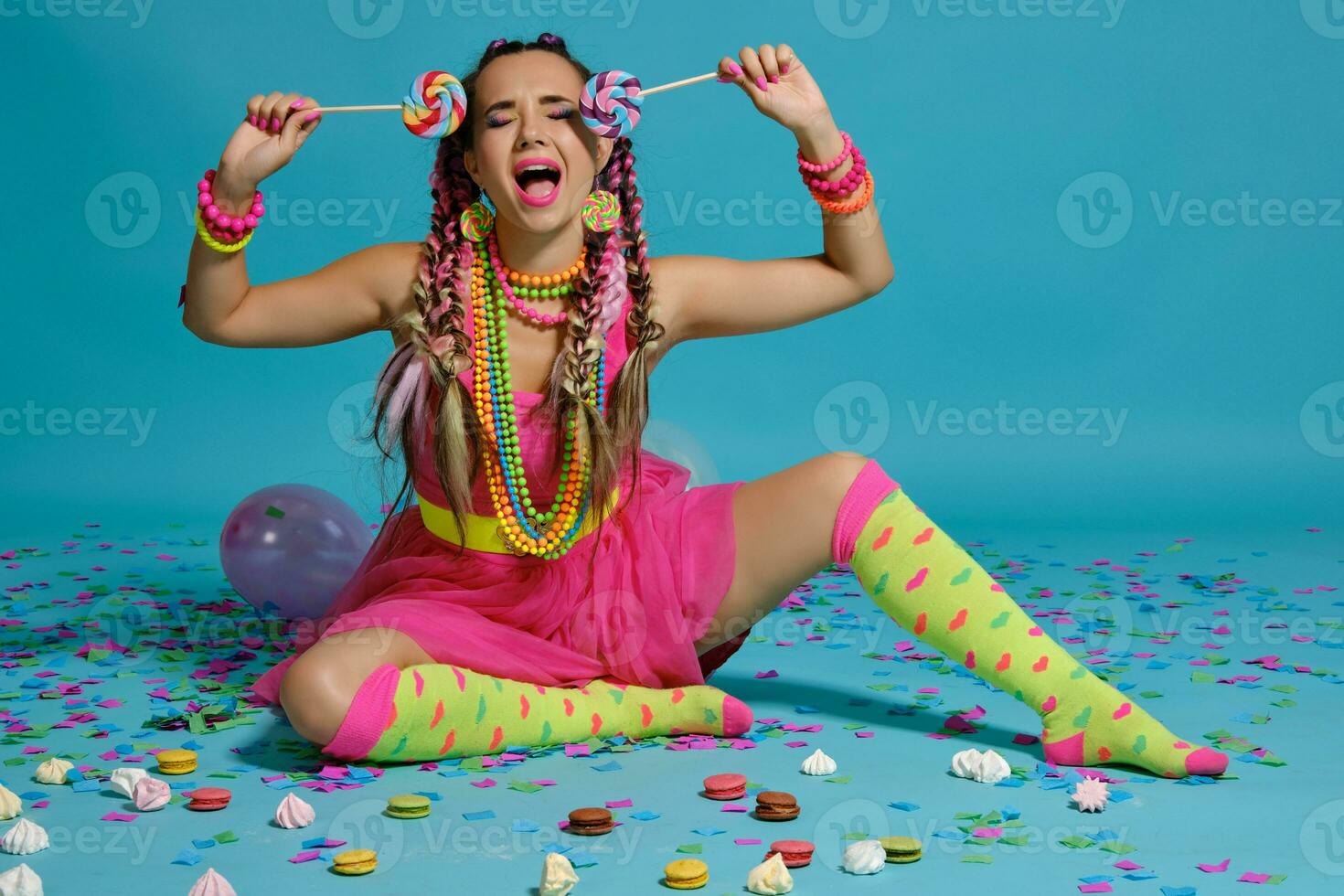Lovely girl with a multi-colored braids hairstyle and bright make-up, posing in studio with lollipop, air balloons and confetti against a blue background. photo