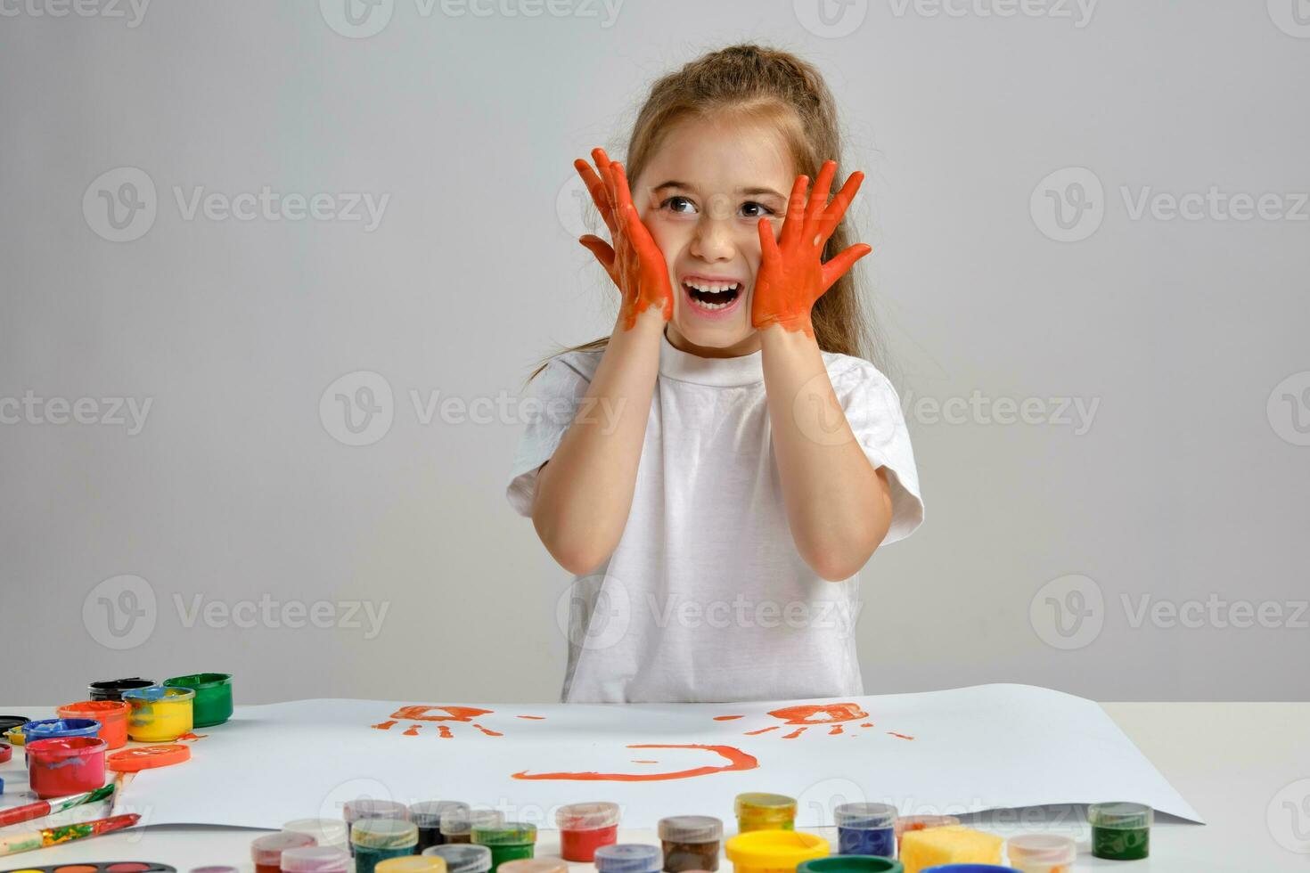 Little girl in white t-shirt sitting at table with whatman and colorful paints on it, painting her cheeks. Isolated on white. Medium close-up. photo