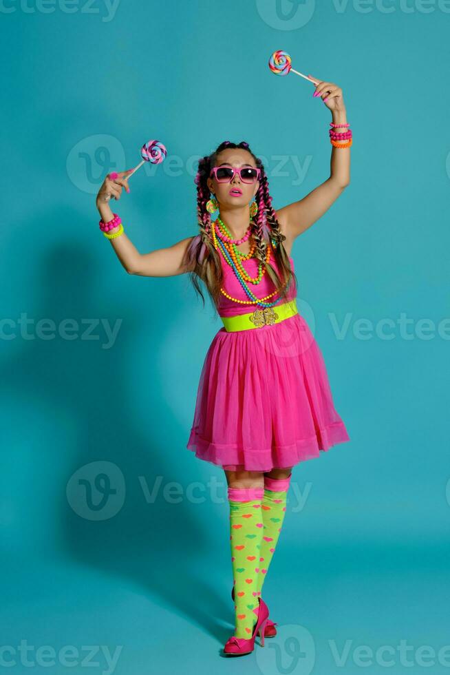 Lovely girl with a multi-colored braids hairstyle and bright make-up, posing in studio against a blue background, holding a lollipop in her hand. photo