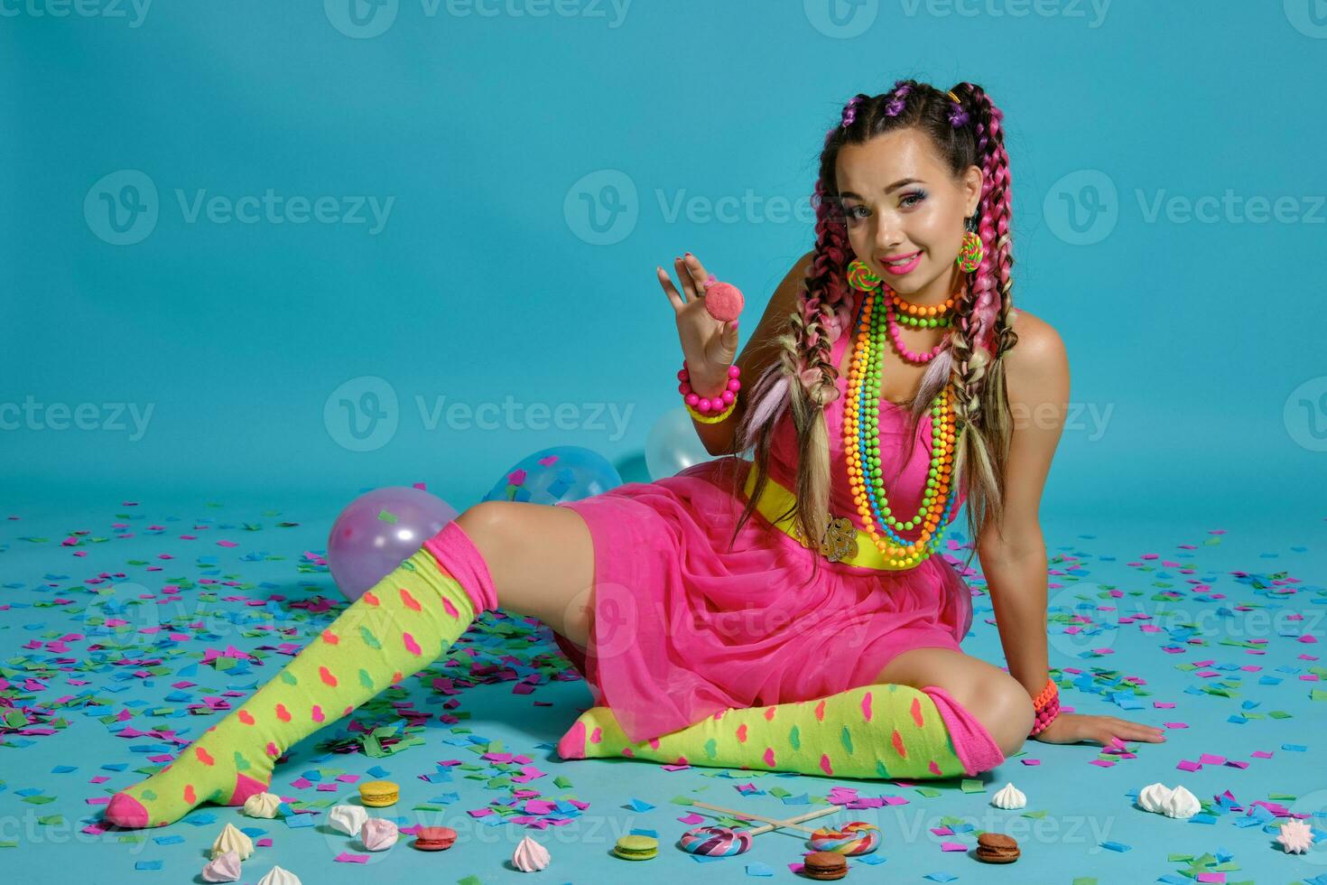 Lovely girl with a multi-colored braids hairstyle and bright make-up, posing in studio with air balloons and confetti against a blue background. photo