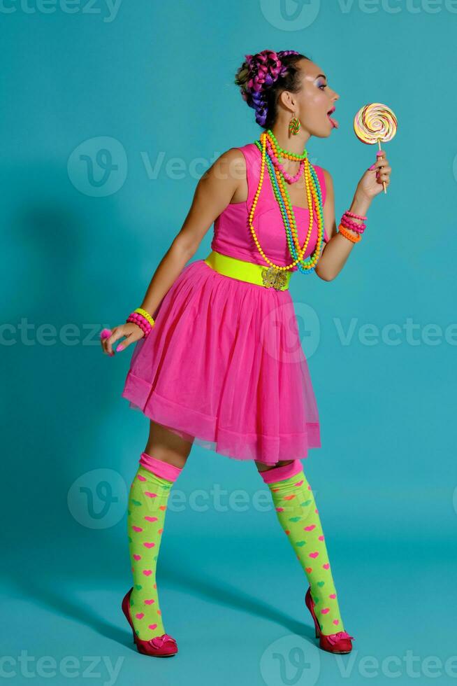 Lovely girl with a multi-colored braids hairstyle and bright make-up, posing in studio against a blue background, holding a lollipop in her hand. photo