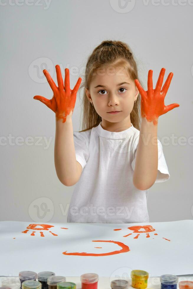 Little girl in white t-shirt sitting at table with whatman and colorful paints, showing her painted hands. Isolated on white. Medium close-up. photo
