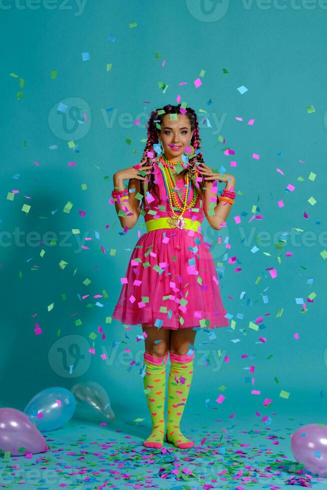 encantador niña con un multicolor trenzas peinado y brillante constituir, posando en estudio con aire globos y papel picado en contra un azul antecedentes. foto