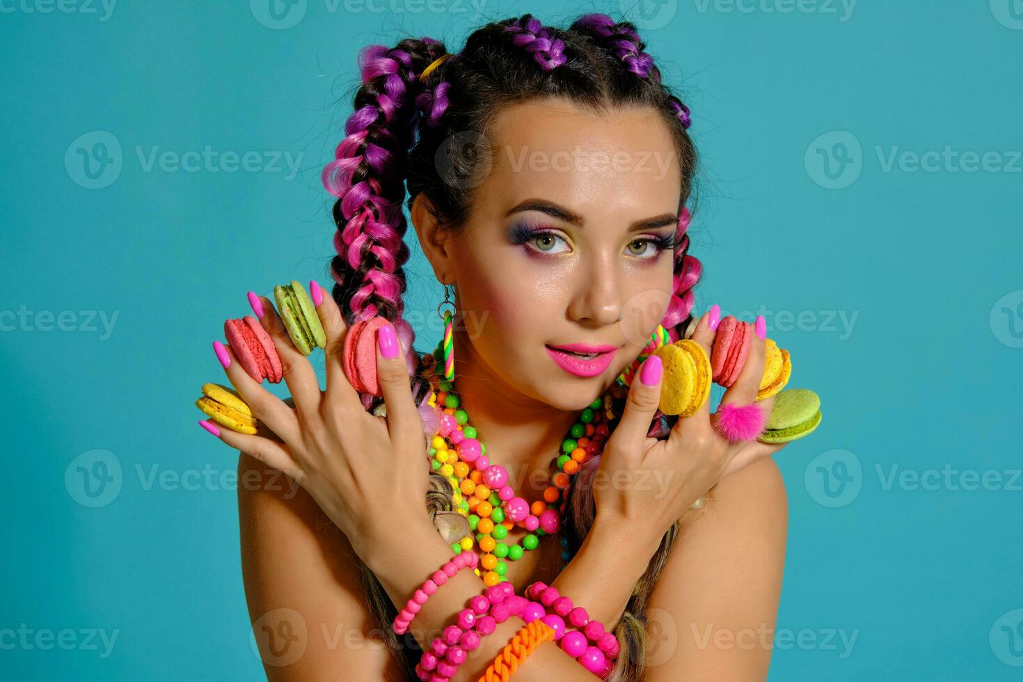 Lovely girl with multi-colored braids hairstyle and bright make-up, holding macarons between her fingers, posing in studio against a blue background. photo