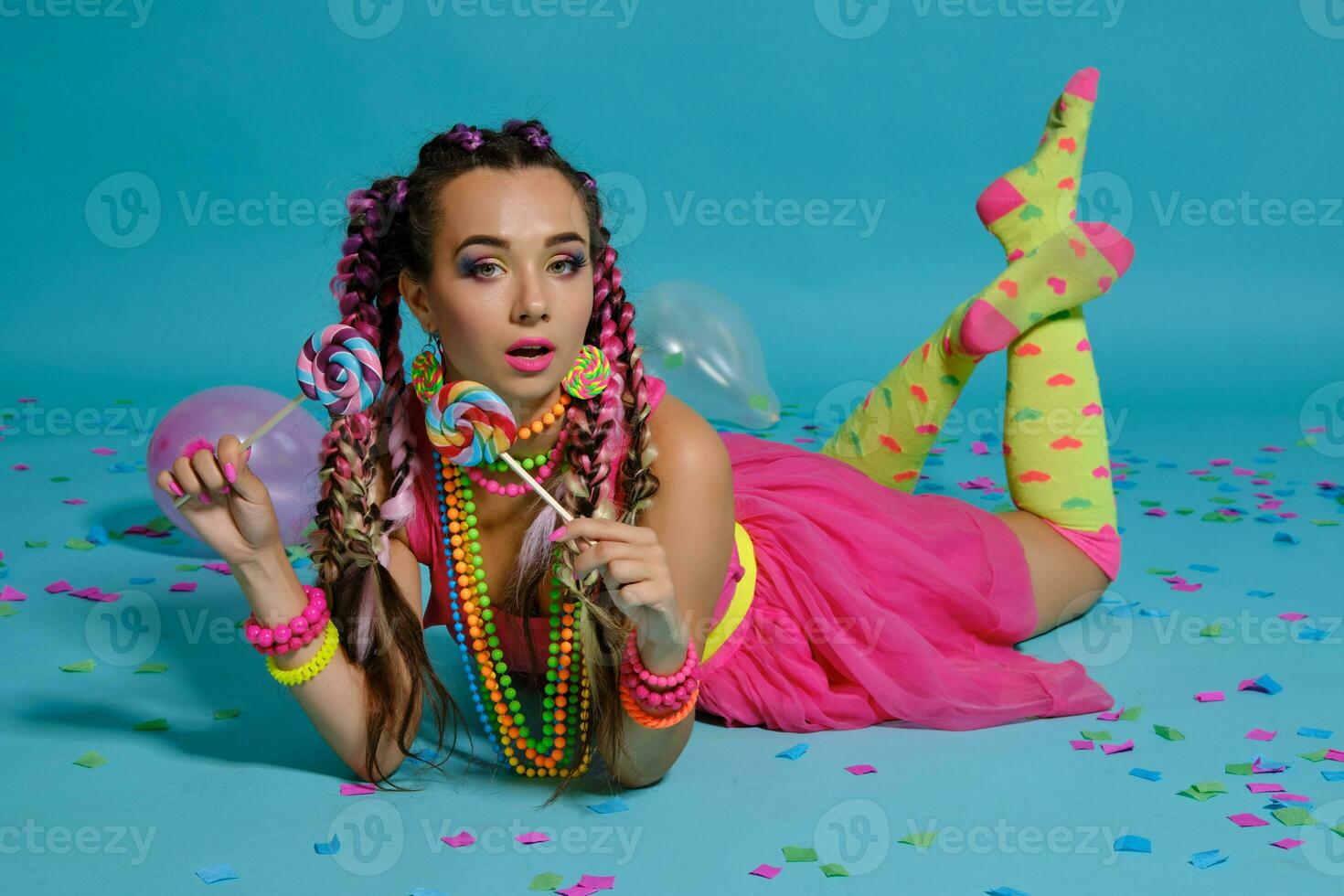 Lovely girl with a multi-colored braids hairstyle and bright make-up, posing in studio with lollipop, air balloons and confetti against a blue background. photo