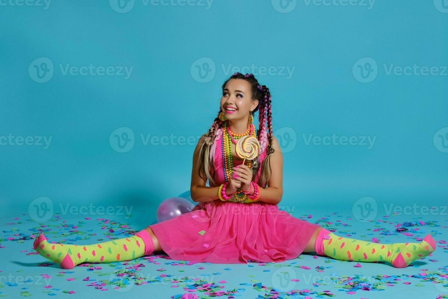 Lovely girl with a multi-colored braids hairstyle and bright make-up, posing in studio with lollipop, air balloons and confetti against a blue background. photo