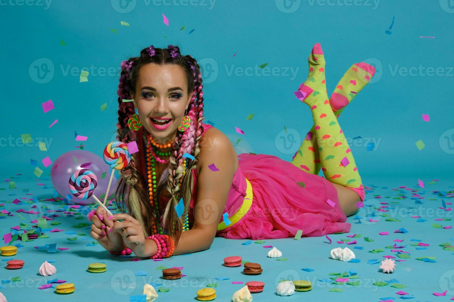 Lovely girl with a multi-colored braids hairstyle and bright make-up, posing in studio with lollipop, air balloons and confetti against a blue background. photo