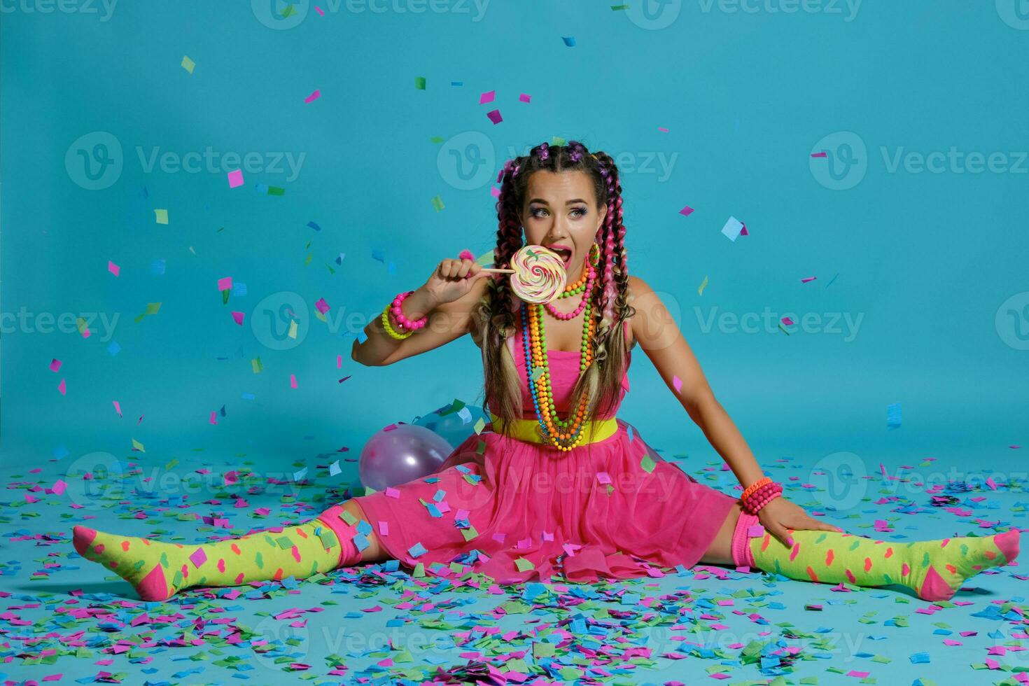 Lovely girl with a multi-colored braids hairstyle and bright make-up, posing in studio with lollipop, air balloons and confetti against a blue background. photo