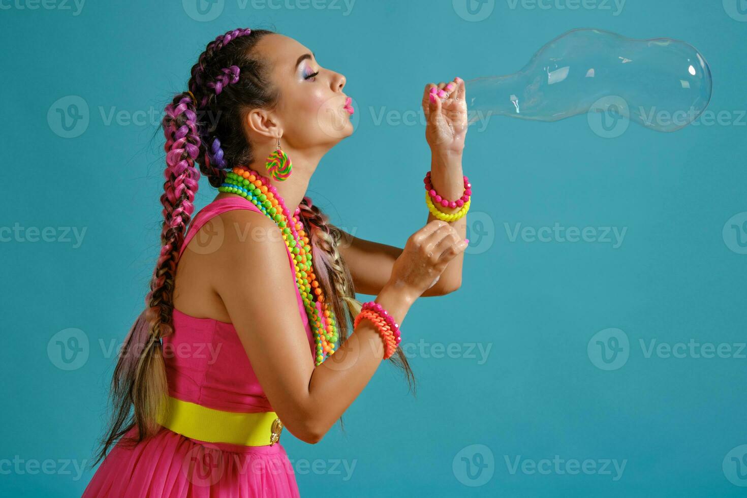 Lovely girl with a multi-colored braids hairstyle and bright make-up, is blowing bubbles using her hands, posing in studio against a blue background. photo