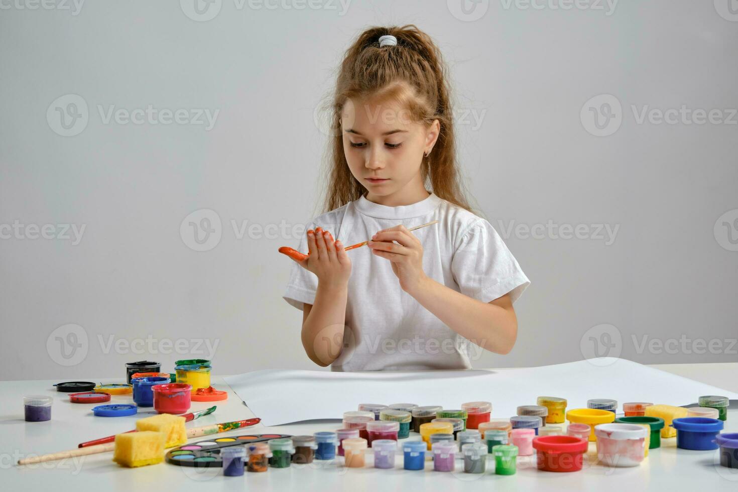 Little girl in white t-shirt sitting at table with whatman and colorful paints on it, painting her hands. Isolated on white. Medium close-up. photo