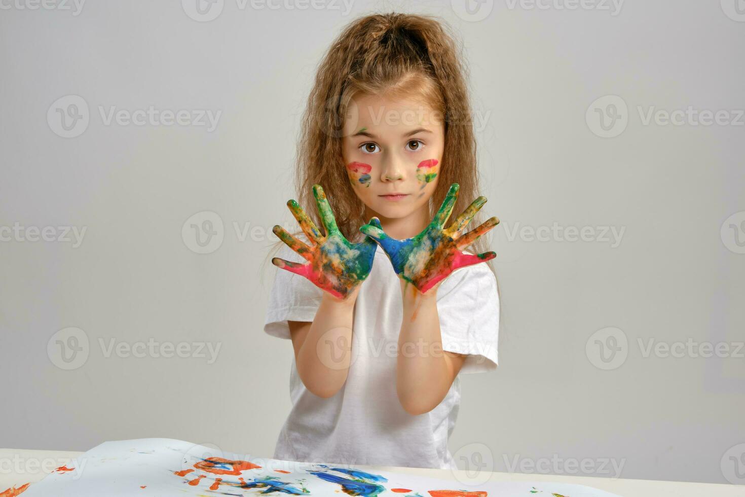 Little girl in white t-shirt sitting at table with whatman and paints on it, posing with painted face and hands. Isolated on white. Medium close-up. photo