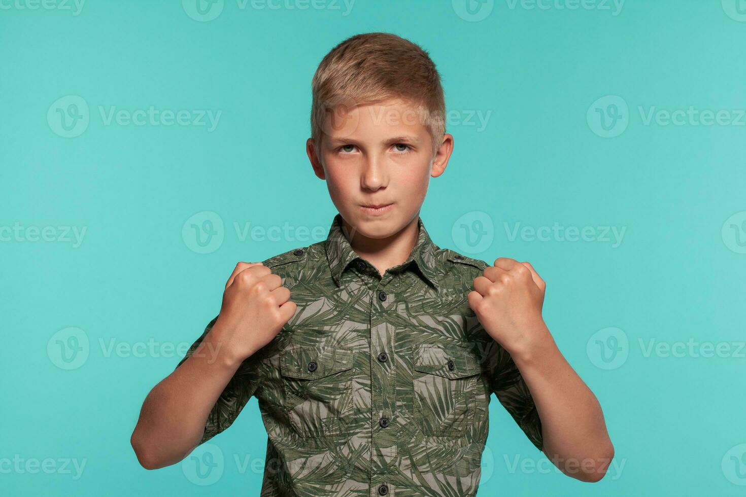 de cerca retrato de un rubia Adolescente chico en un verde camisa con palma impresión posando en contra un azul estudio antecedentes. concepto de sincero emociones foto