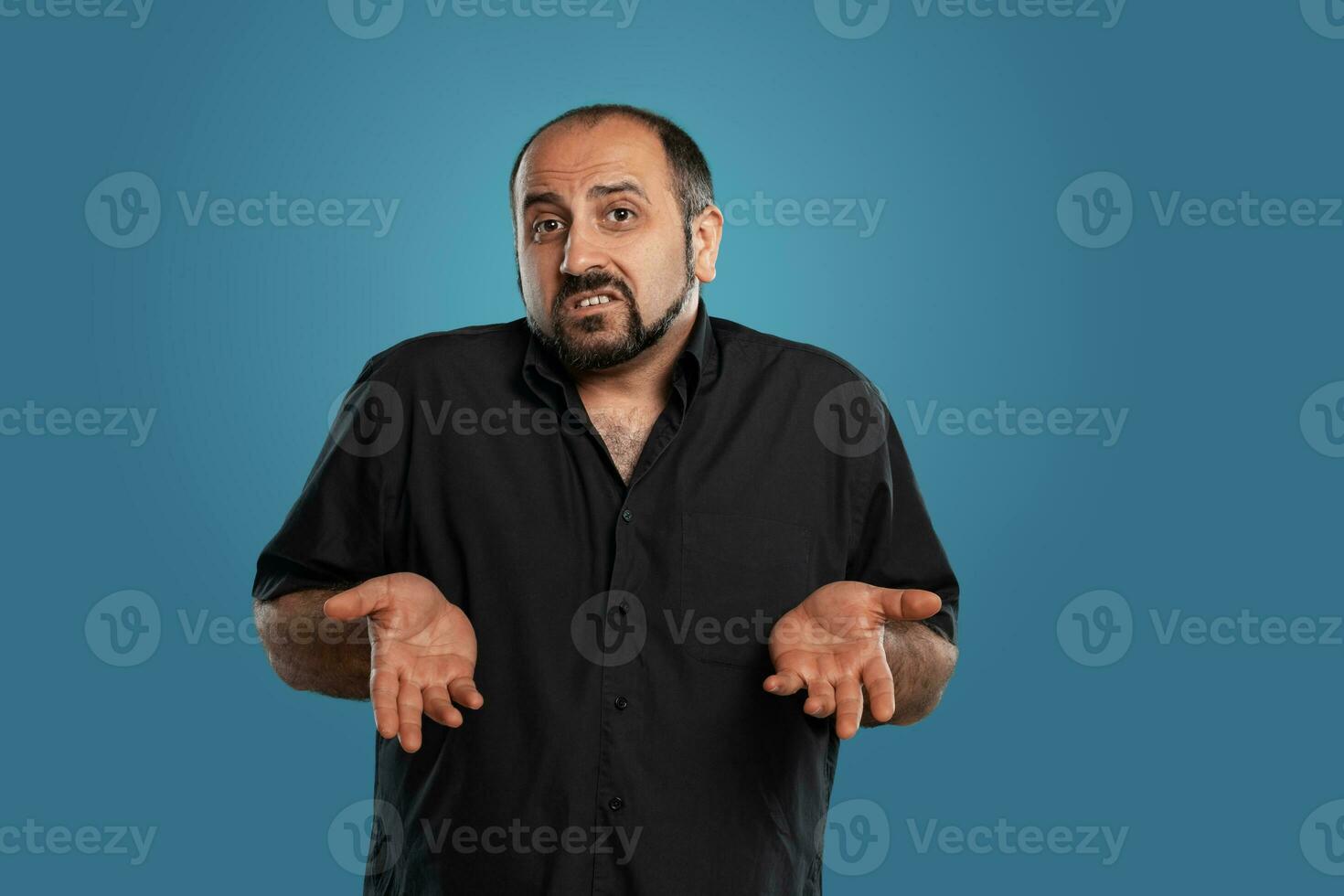 de cerca retrato de un moreno de edad mediana hombre con barba, vestido en un negro camiseta y posando en contra un azul antecedentes. foto