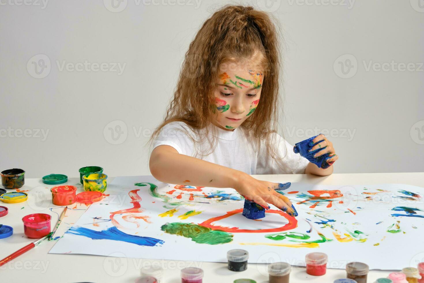 pequeño niña en blanco camiseta sentado a mesa con qué hombre y pinturas, dibujo en él, posando con pintado rostro, manos. aislado en blanco. de cerca. foto