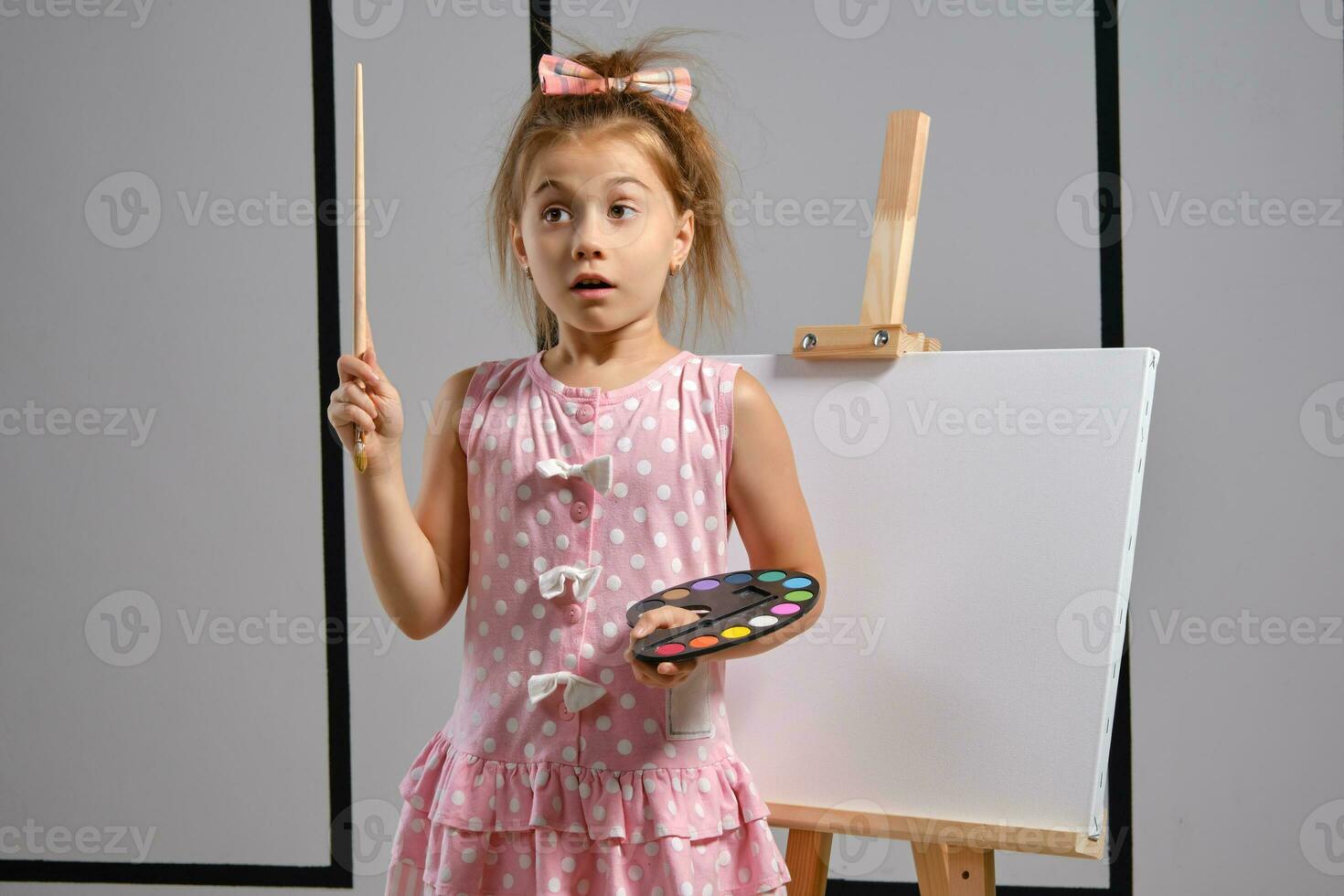 Little girl artist in a pink dress is standing behind easel and painting with brush on canvas at art studio with white walls. Medium close-up shot. photo