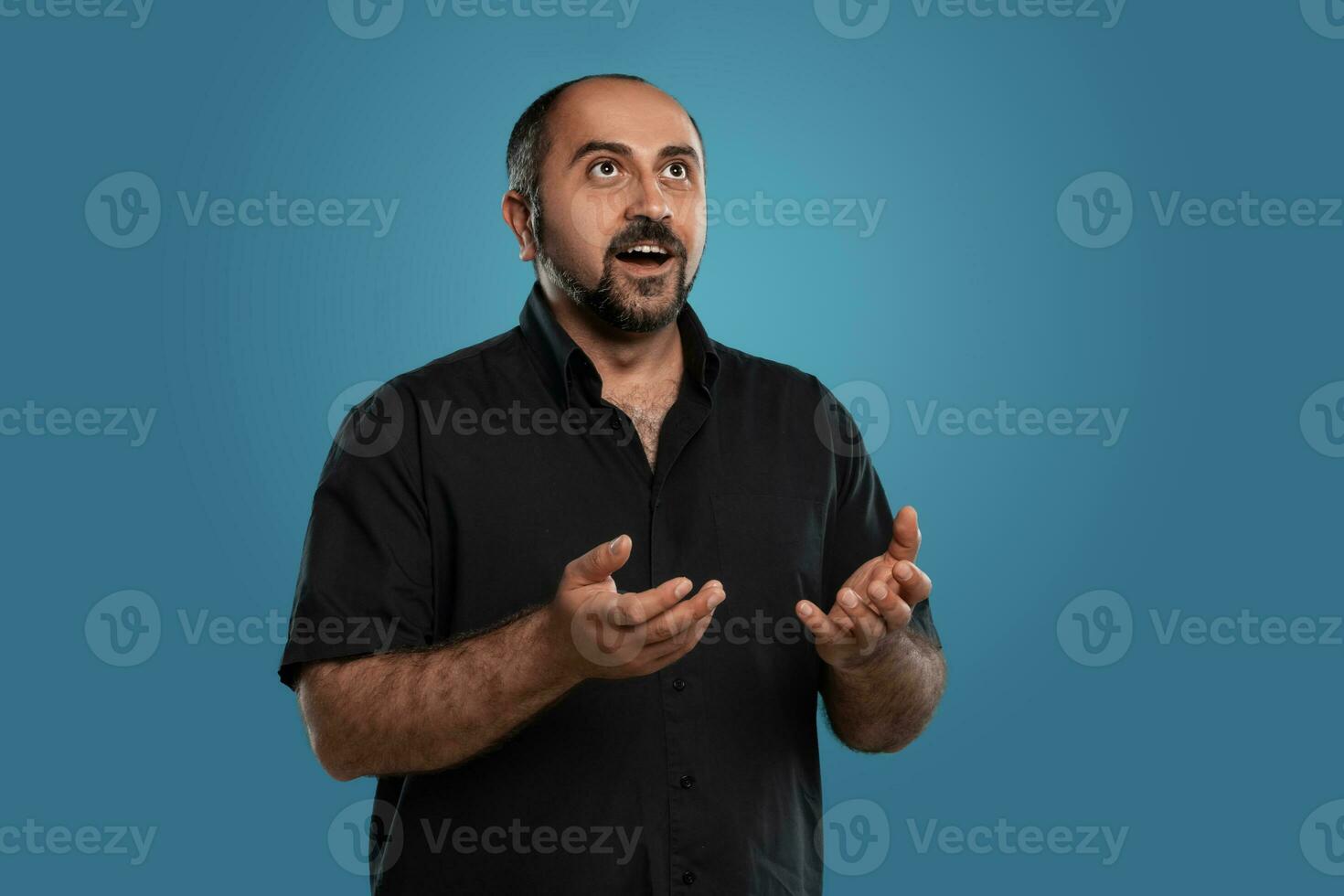 de cerca retrato de un moreno de edad mediana hombre con barba, vestido en un negro camiseta y posando en contra un azul antecedentes. foto