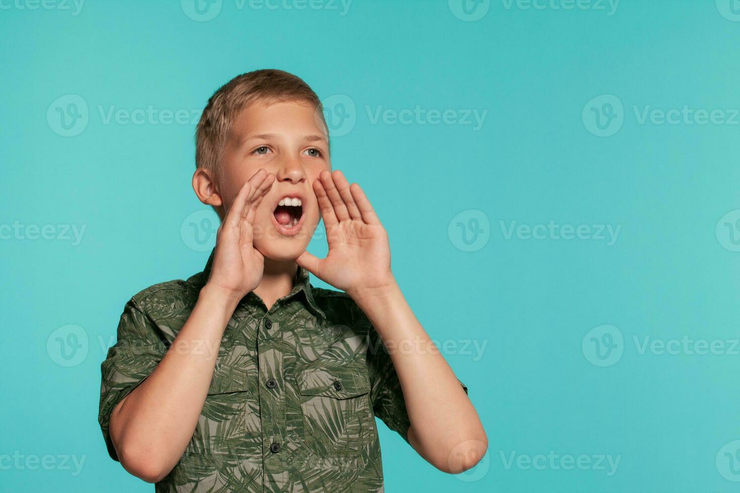 Close-up portrait of a blonde teenage boy in a green shirt with palm print posing against a blue studio background. Concept of sincere emotions. photo