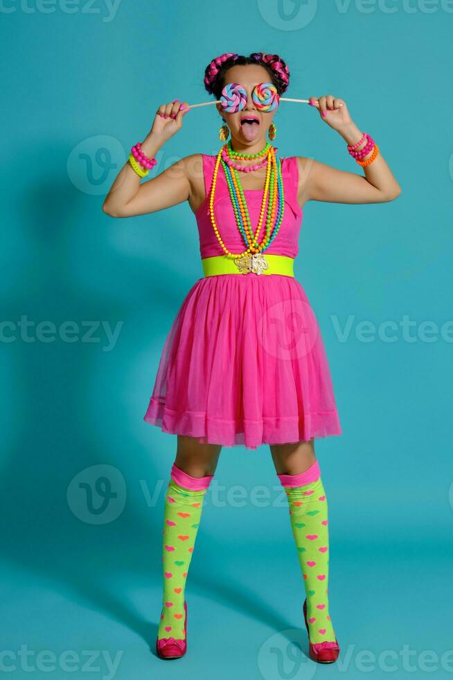 Lovely girl with a multi-colored braids hairstyle and bright make-up, posing in studio against a blue background, holding a lollipop in her hand. photo