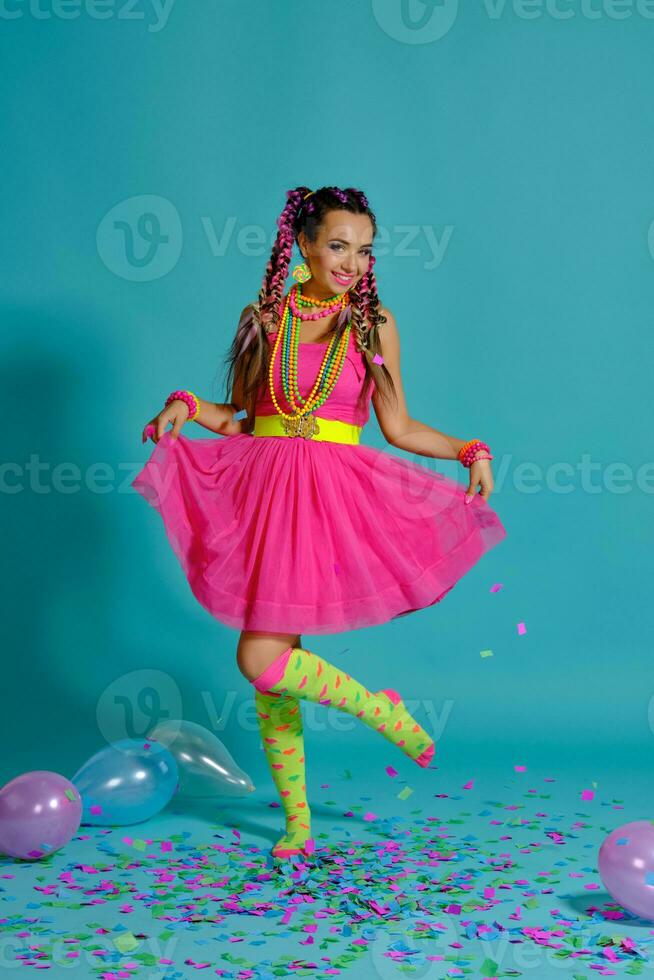 encantador niña con un multicolor trenzas peinado y brillante constituir, posando en estudio con aire globos y papel picado en contra un azul antecedentes. foto