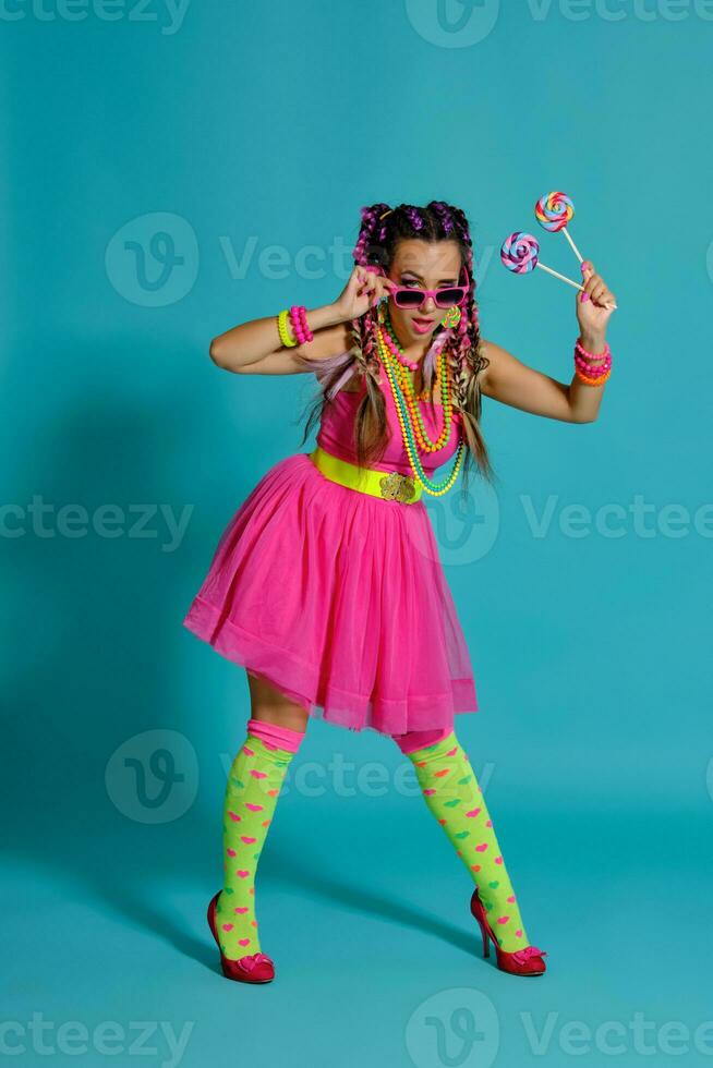 encantador niña con un multicolor trenzas peinado y brillante constituir, posando en estudio en contra un azul fondo, participación un pirulí en su mano. foto