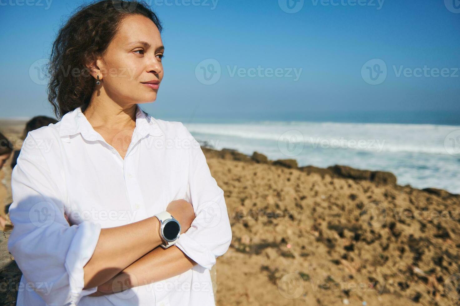 joven adulto mujer con brazos doblada en pie terminado marina fondo, disfruta hermosa soleado día, pensativamente mirando lejos foto