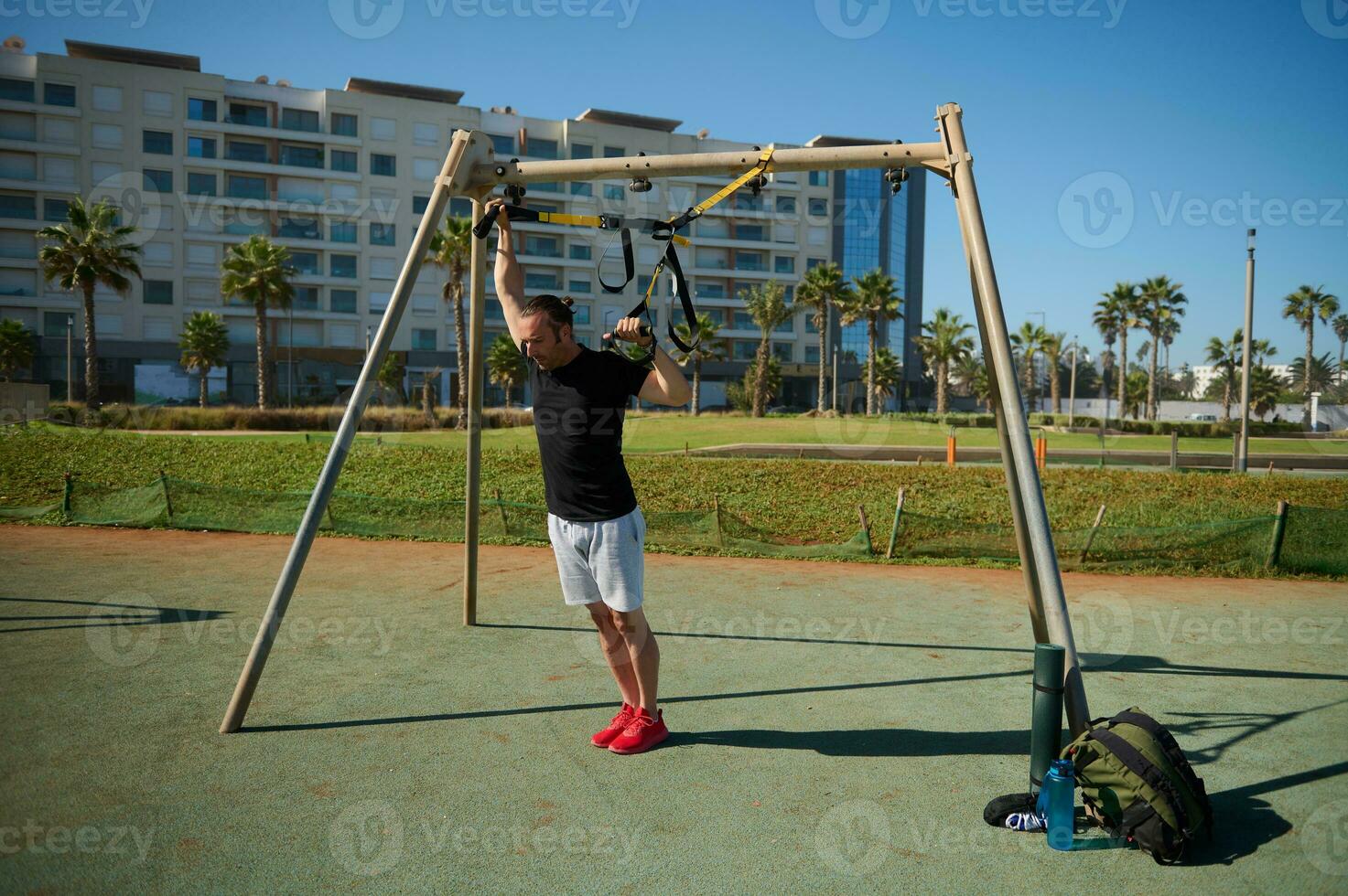Young adult athletic guy training arms using suspension fitness straps during an outdoor workout in urban sports ground photo