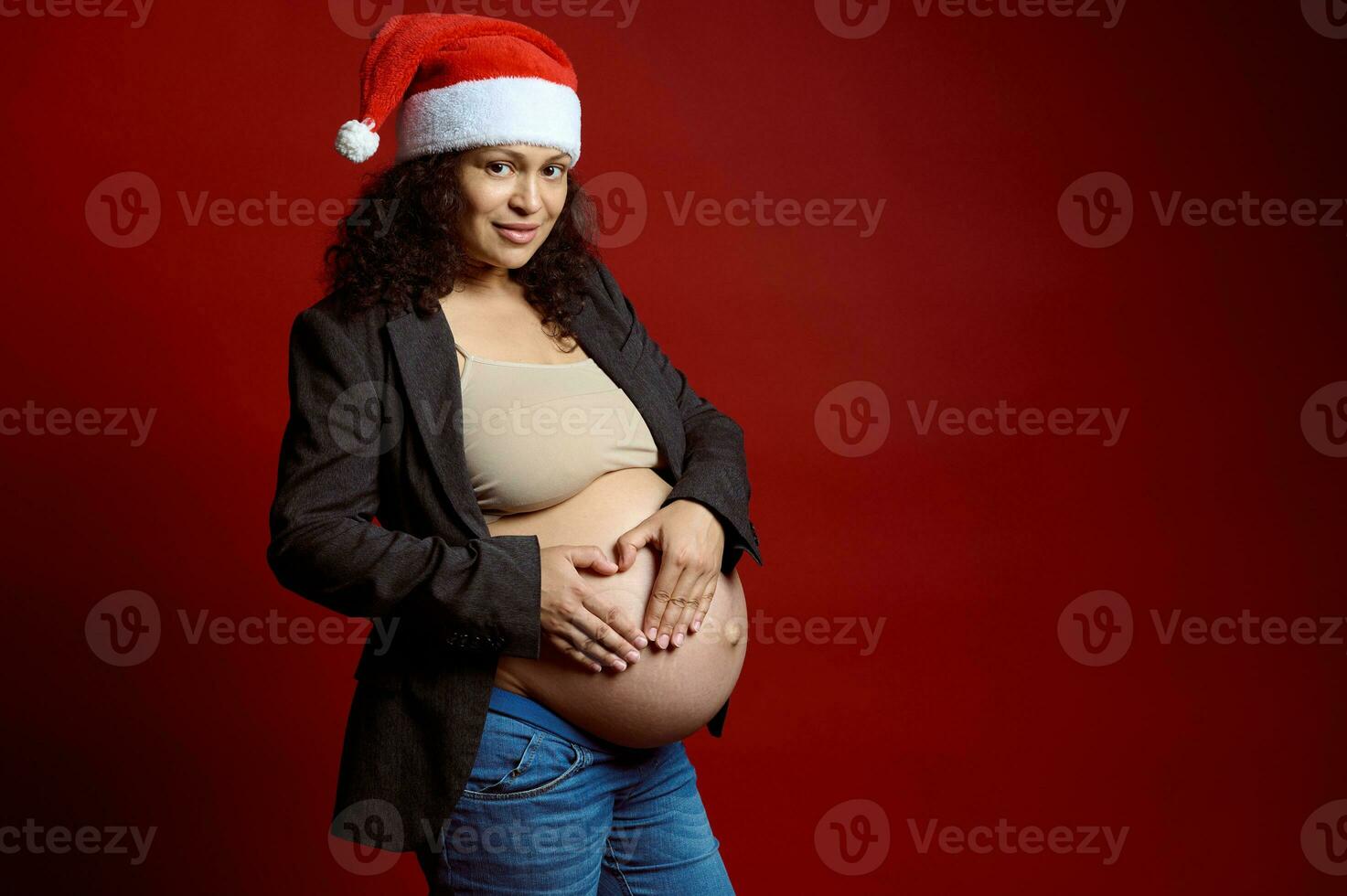 Charming pregnant woman in Santa hat, forming a shape of heart over her naked belly, smiling at camera, red background photo