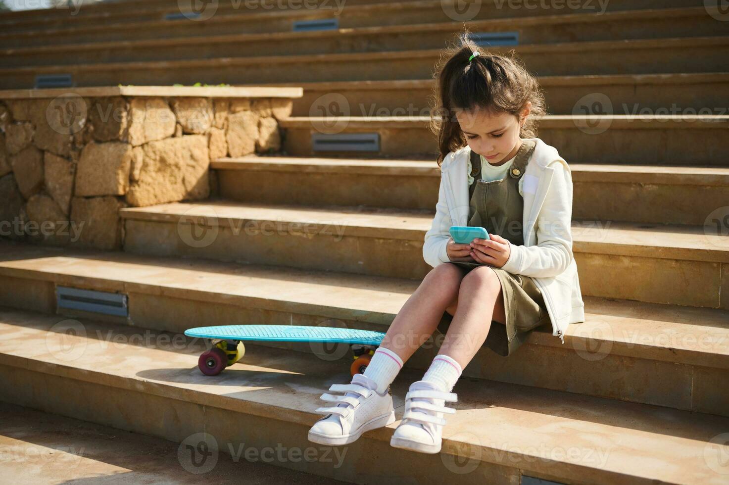 Adorable child girl using mobile phone, browses internet, plays online video games, watches cartoons, sitting on steps photo