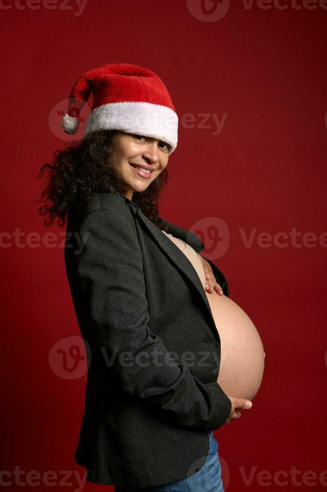 Happy young adult pregnant woman, expectant mother wearing Santa hat, holding her belly, confidently looking at camera photo