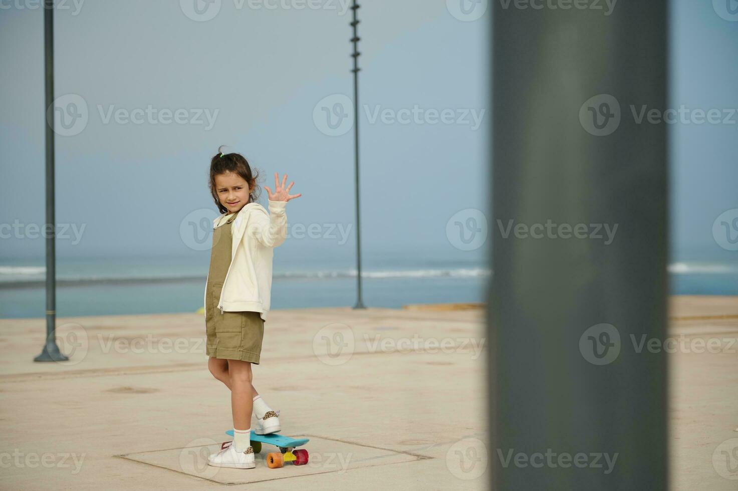 Adorable little child girl greeting with hand, standing on her blue skateboard on one leg on a modern urban skate park photo