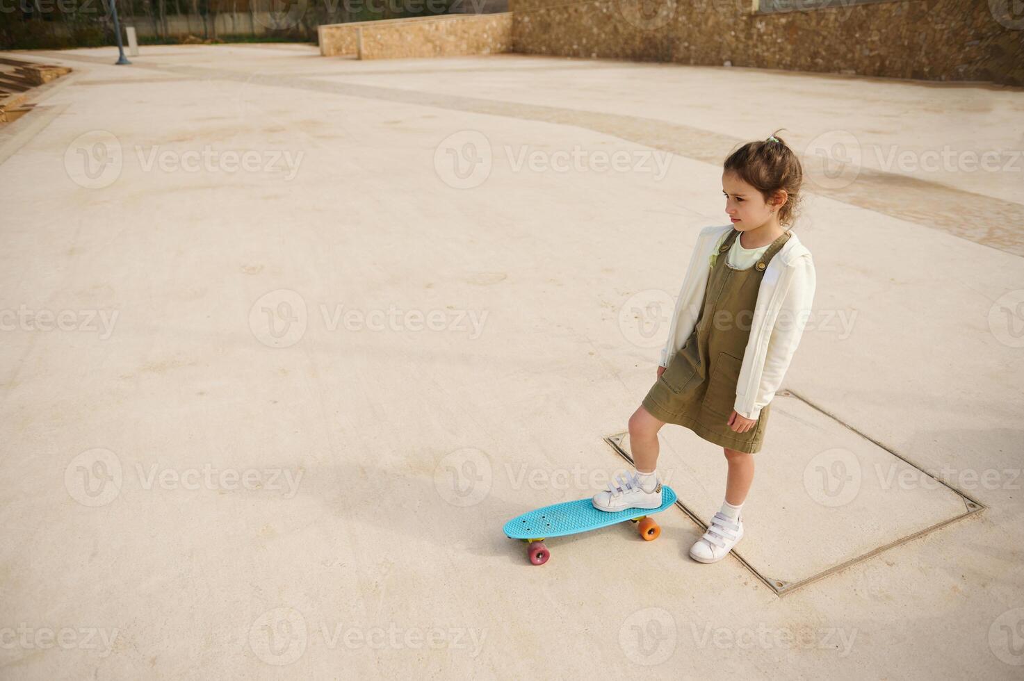 View from above of a beautiful little girl standing on her skateboard on one leg, dreamily looking into the distance photo
