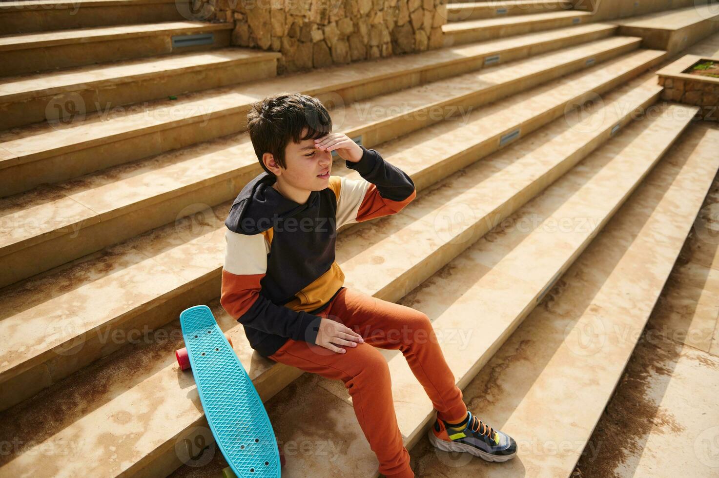 Teenage student boy, schoolboy in sportswear sitting on steps in the urban skatepark, thoughtfully looking into distance photo