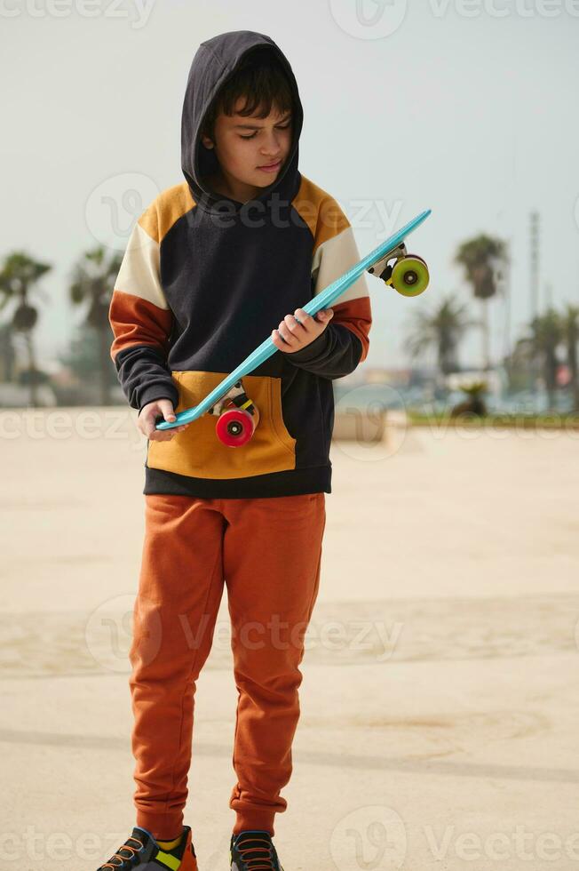 Caucasian teenage boy dressed in stylish sportswear, holding skateboard and looking down, standing at outdoor skatepark photo