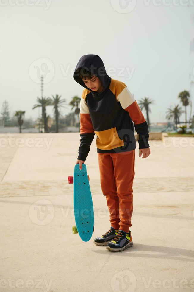 Full length portrait of stylish schoolboy in sportswear, holding skateboard, standing on an outdoor skatepark playground photo