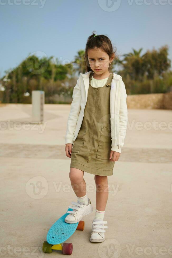 Upset Caucasian child girl standing on her skateboard on one leg on skatepark, expressing sad emotions looking at camera photo