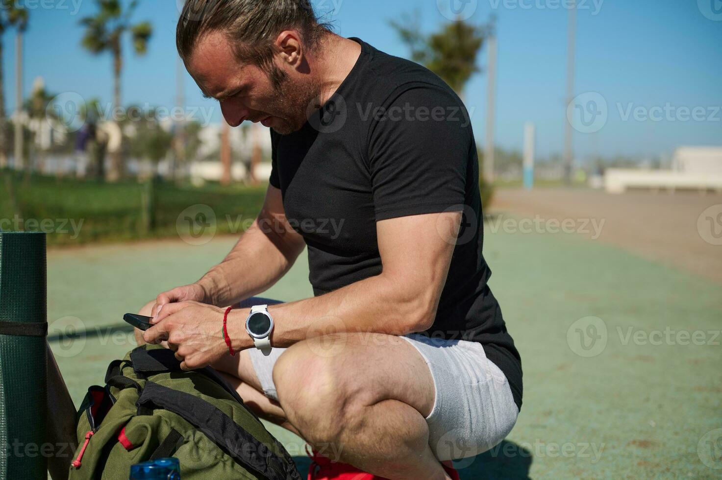 Handsome sportsman checking his mobile phone, sitting near backpack on the sportsground. People. Active lifestyle. Sport photo