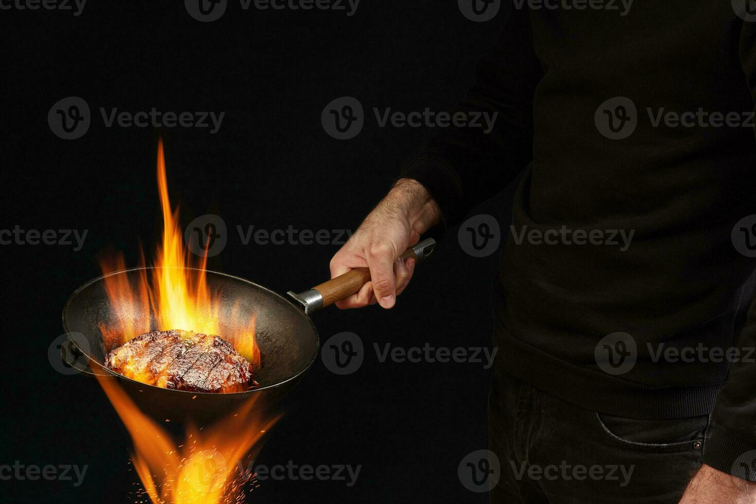 Young fellow dressed in jumper and jeans. He is holding burning wok pan above fire and frying beef steak, against black studio background. Side view photo