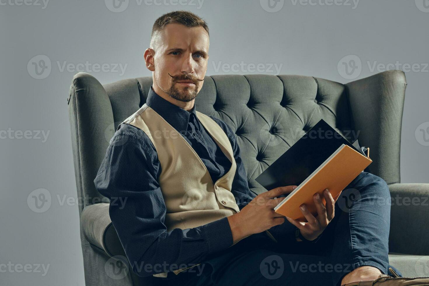 hombre con elegante Bigote, vestido en negro camisa y pantalones, beige chaleco es sentado en oscuro sofá, leyendo un revista. gris fondo, de cerca. foto