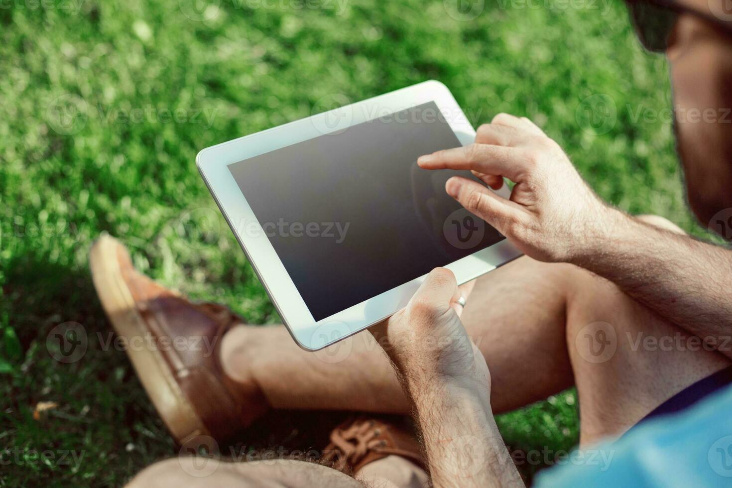 Close-up of casual dressed young man using modern digital tablet while sitting at the park, sun light photo
