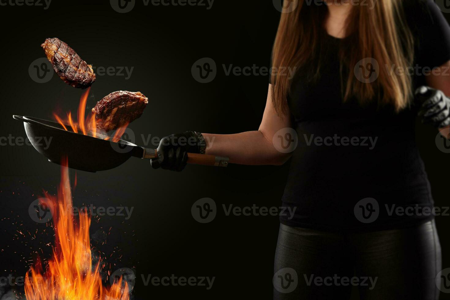 Cook with tattooed hands, dressed in leggings and t-shirt. Holding wok pan above fire and frying two beef steaks against black background. Side view photo