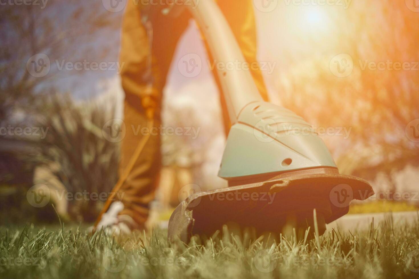 Male in casual outfit is mowing green grass with handheld electric lawn mower on his backyard. Gardening care tools and services. Close up photo