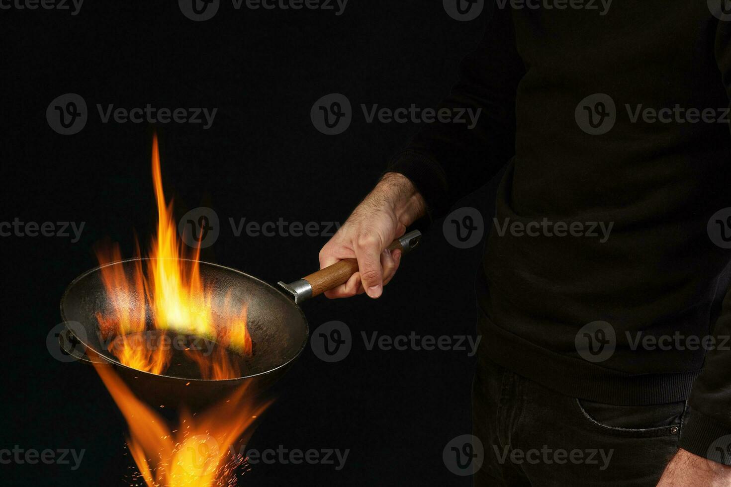 Young guy dressed in jumper and jeans. He is holding burning wok pan above fire, against black studio background. Cooking concept. Close up photo