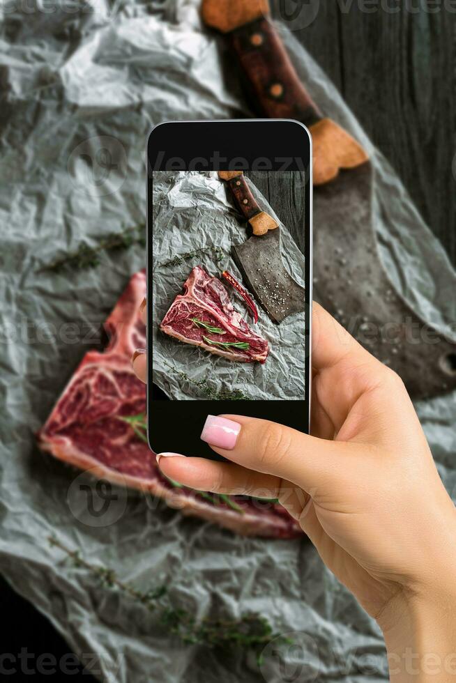 Photographing food concept - woman takes picture of raw dry aged t-bone steaks for grill with fresh herbs and cleaver photo