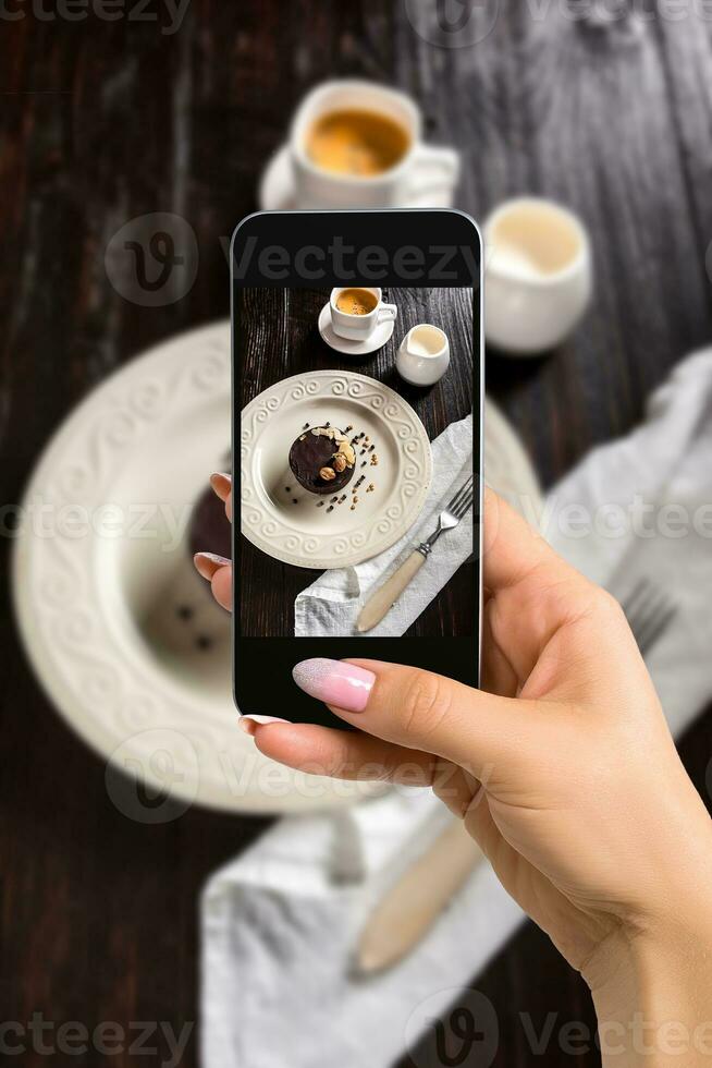 Photographing food concept - woman takes picture of beautiful decorated chocolate cake with golden nuts and a cup of coffee photo