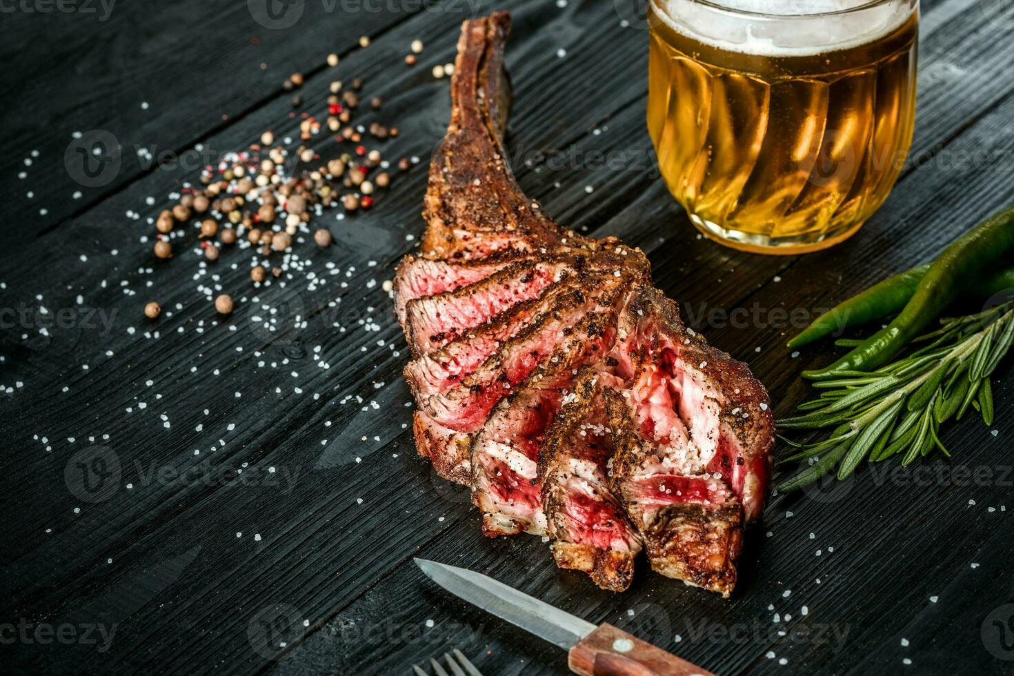 Barbecue dry aged rib of beef with spice, vegetables and a glass of light beer close-up on black wooden background photo